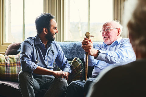 Male doctor with an older man with a walking aid. Both sitting down on the sofa smiling.